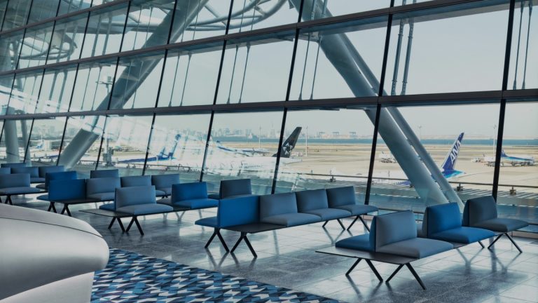 Modern airport terminal with large glass windows overlooking parked airplanes. Empty blue seating areas create a calm, spacious atmosphere.